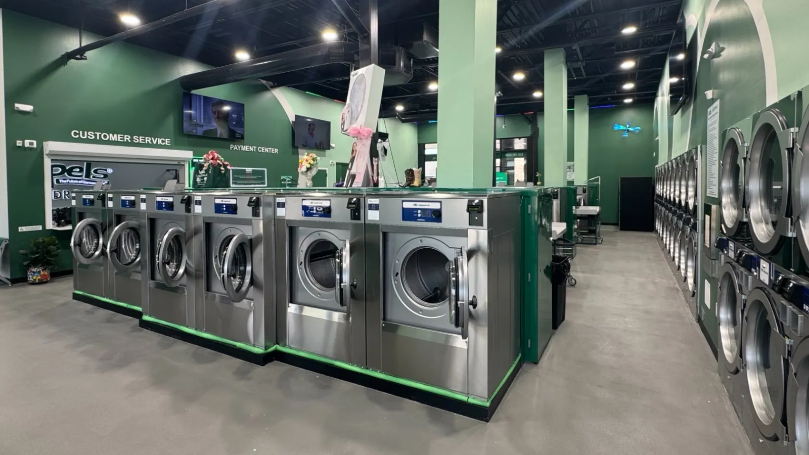 Modern laundromat interior featuring rows of stainless steel washing machines and dryers with green walls.