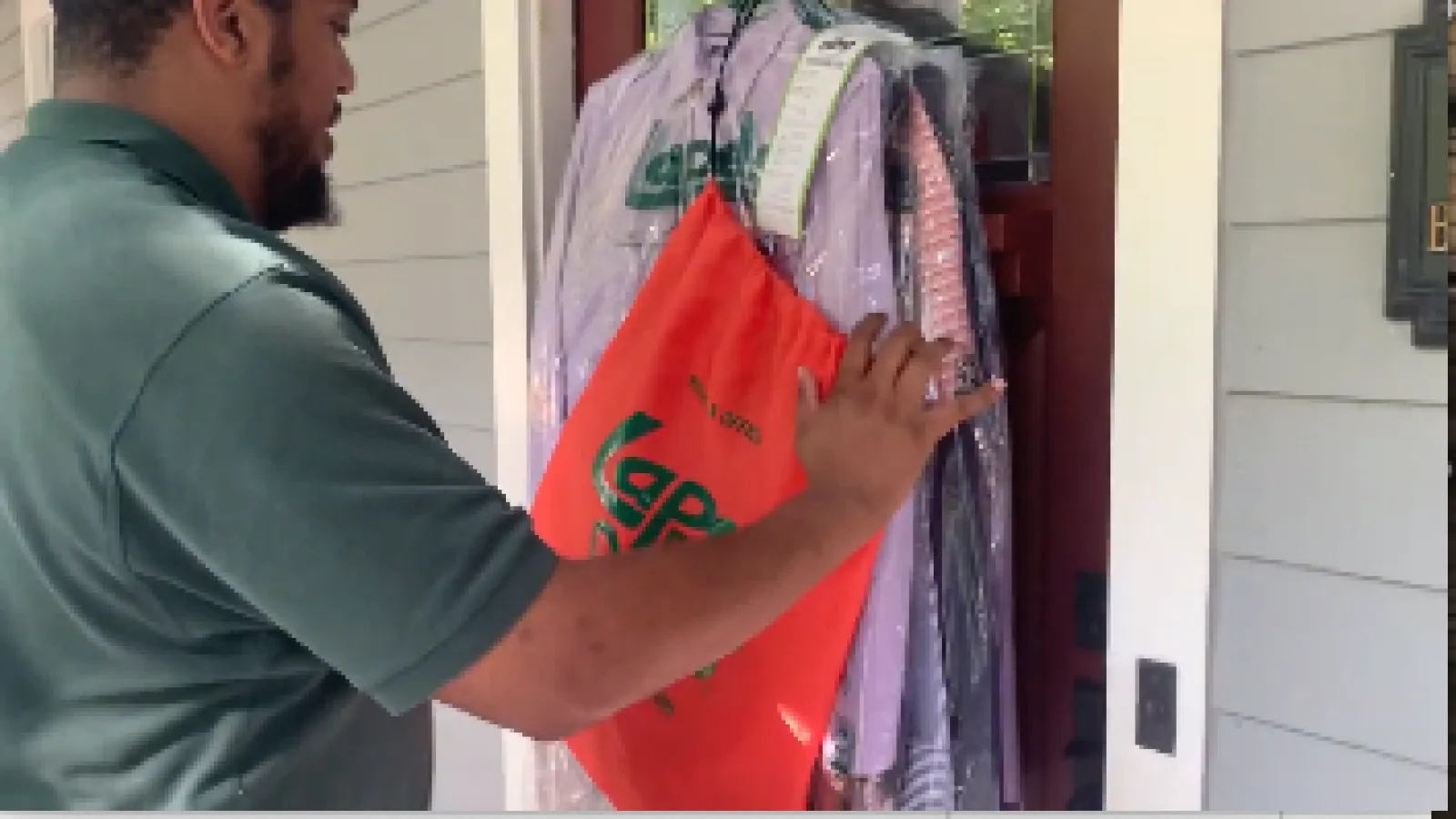 Man delivering dry cleaning with clothes on hangers and branded red bag at a house entrance