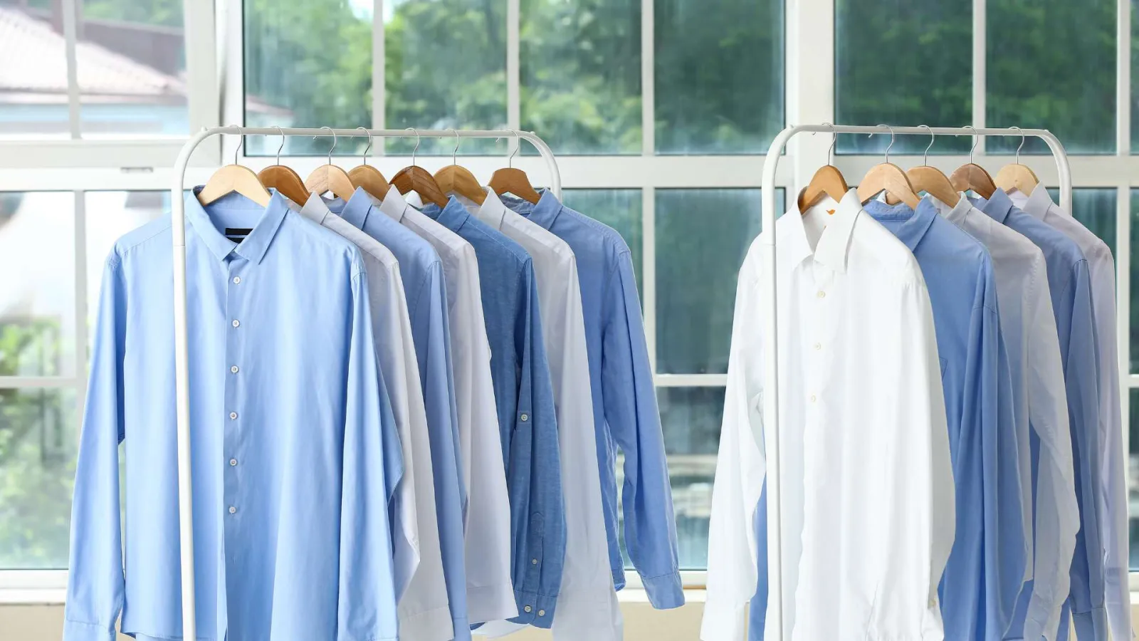Row of blue and white dress shirts hanging on racks in front of large windows with natural light.