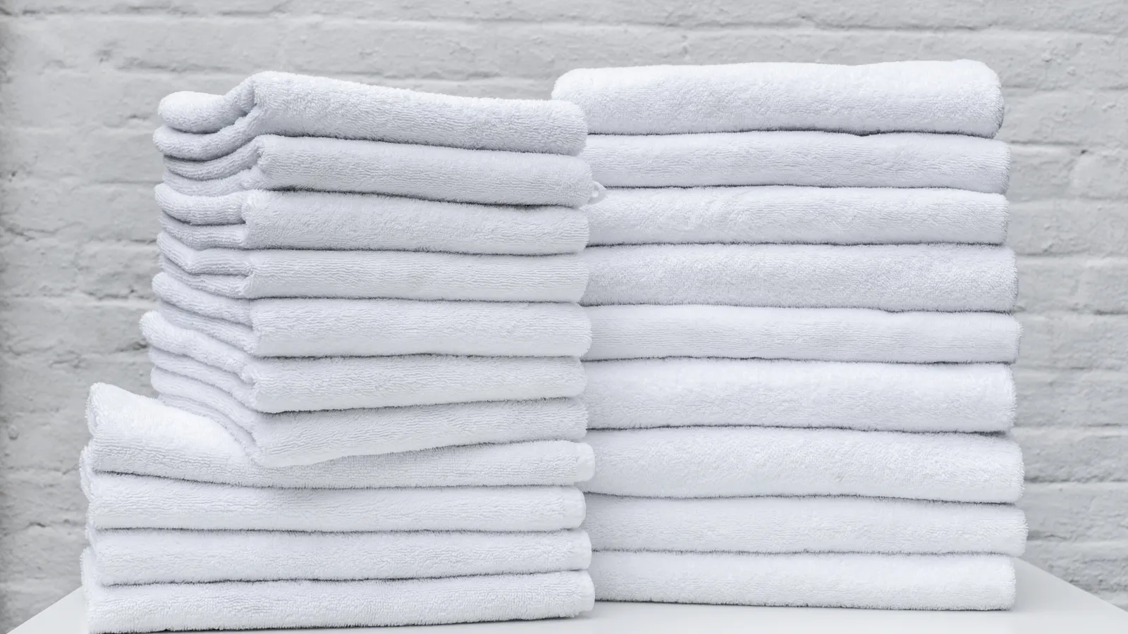 Stacks of neatly folded white towels arranged on a white surface against a gray brick wall.