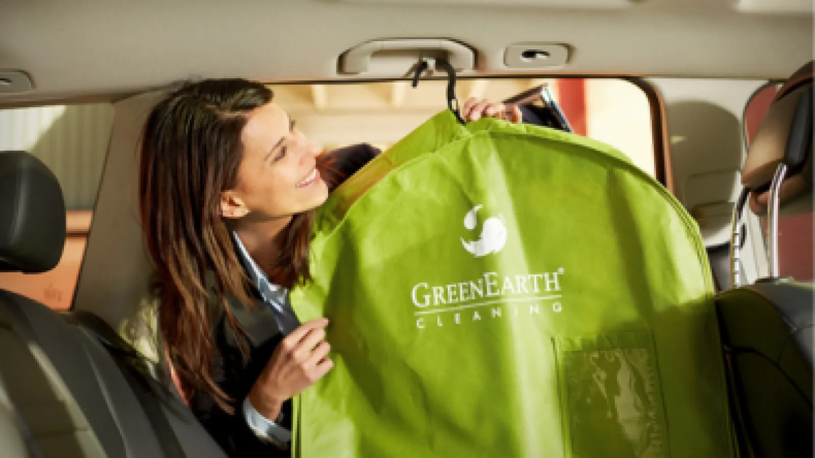 Smiling woman inside car holding a green garment bag with GreenEarth Cleaning logo on it.