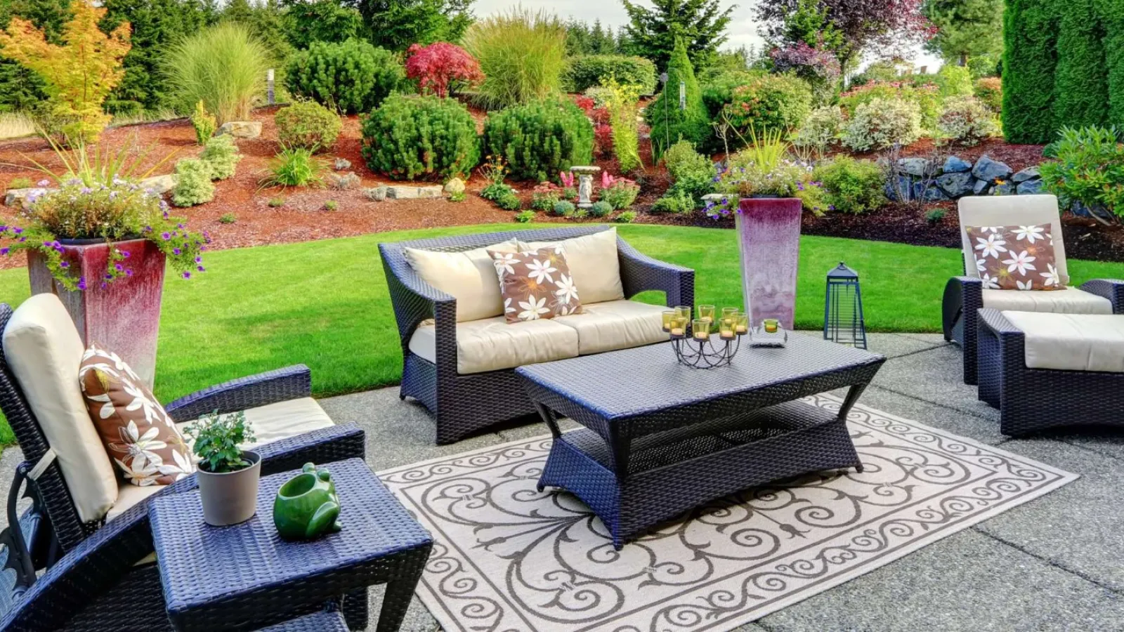 Modern outdoor patio set with chairs, sofa, coffee table on rug surrounded by lush garden and blue sky.