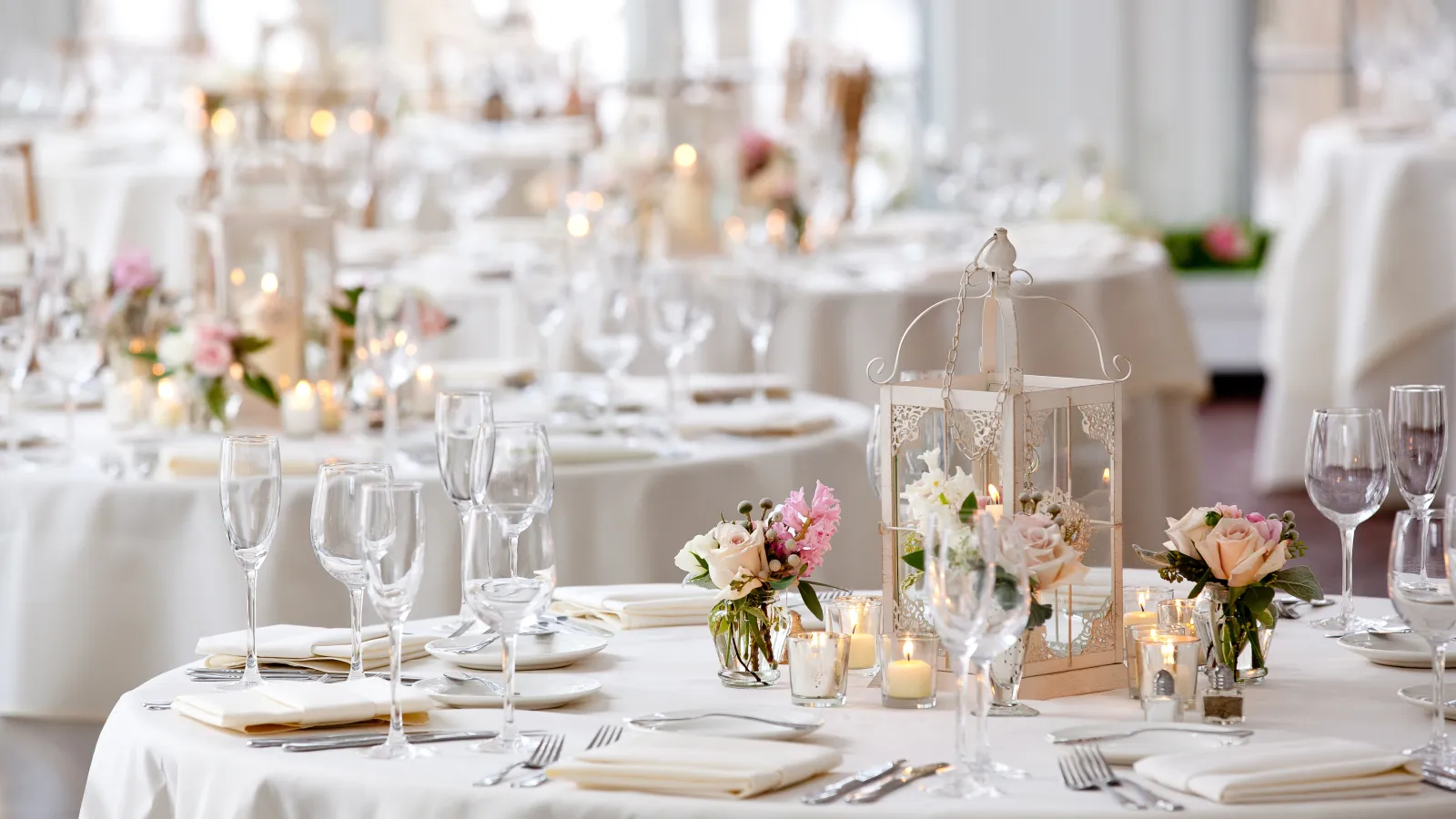 Elegantly set round tables with white linens, floral centerpieces, candles, and glassware at a wedding reception.