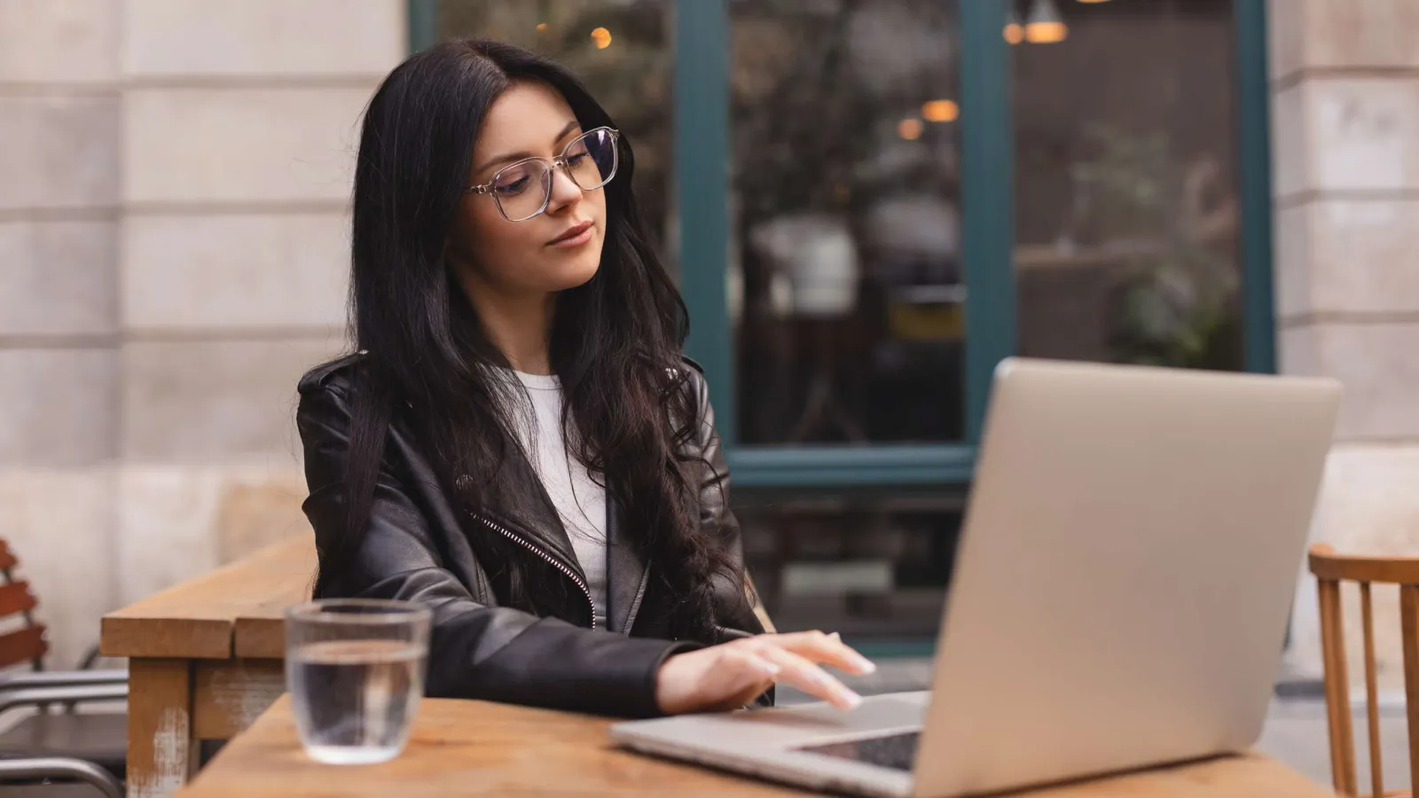 Young woman with glasses working on a laptop at an outdoor wooden table with a glass of water.