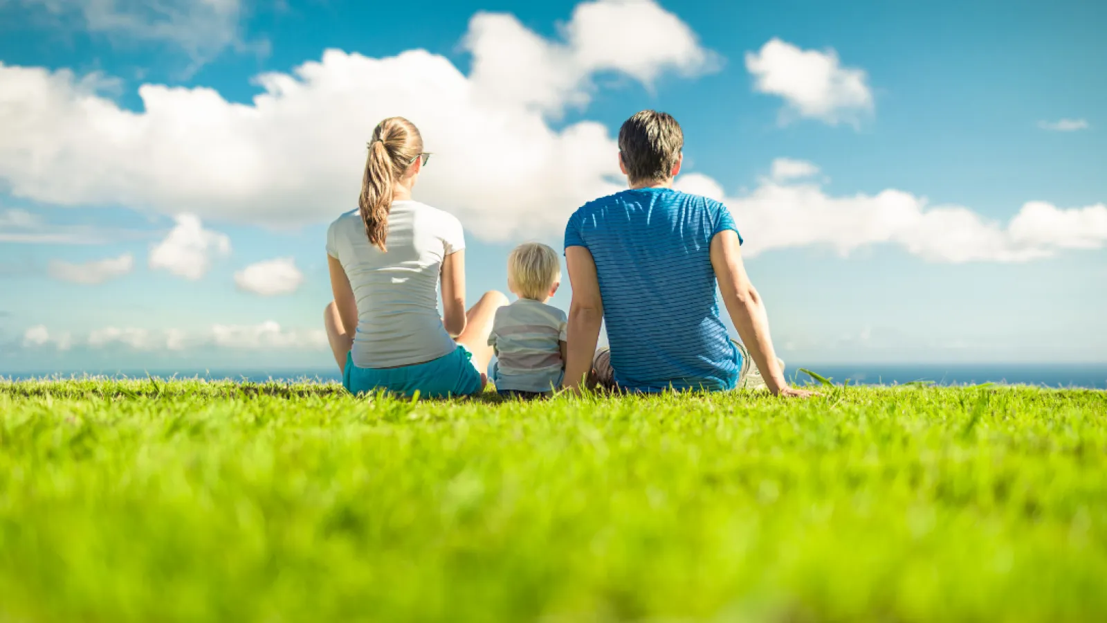 Family of three sitting on grassy hill under blue sky with white clouds, enjoying outdoor view together.