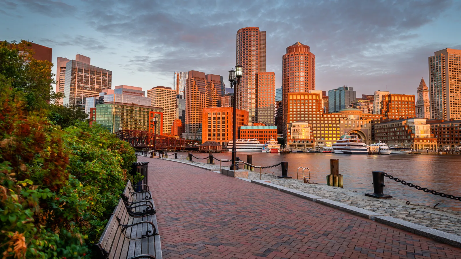 Sunset view of Boston skyline with waterfront promenade, benches, and boats along the harbor.