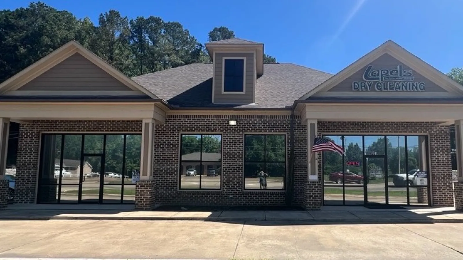 Modern brick dry cleaning storefront under clear blue sky with an American flag and green lawn in front.