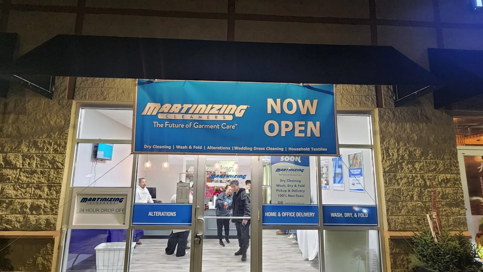 Martinizing Cleaners storefront at night with blue banners announcing now open and laundry services available.