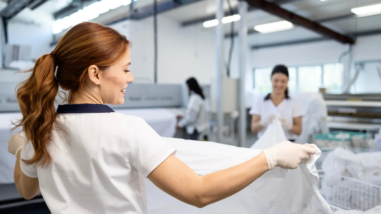 Smiling woman folding white laundry in a bright, industrial laundry facility with colleagues working in background