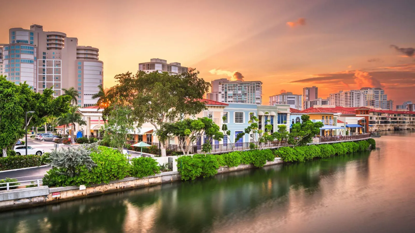 Sunset over waterfront homes and high-rise buildings with lush greenery and calm water reflections.