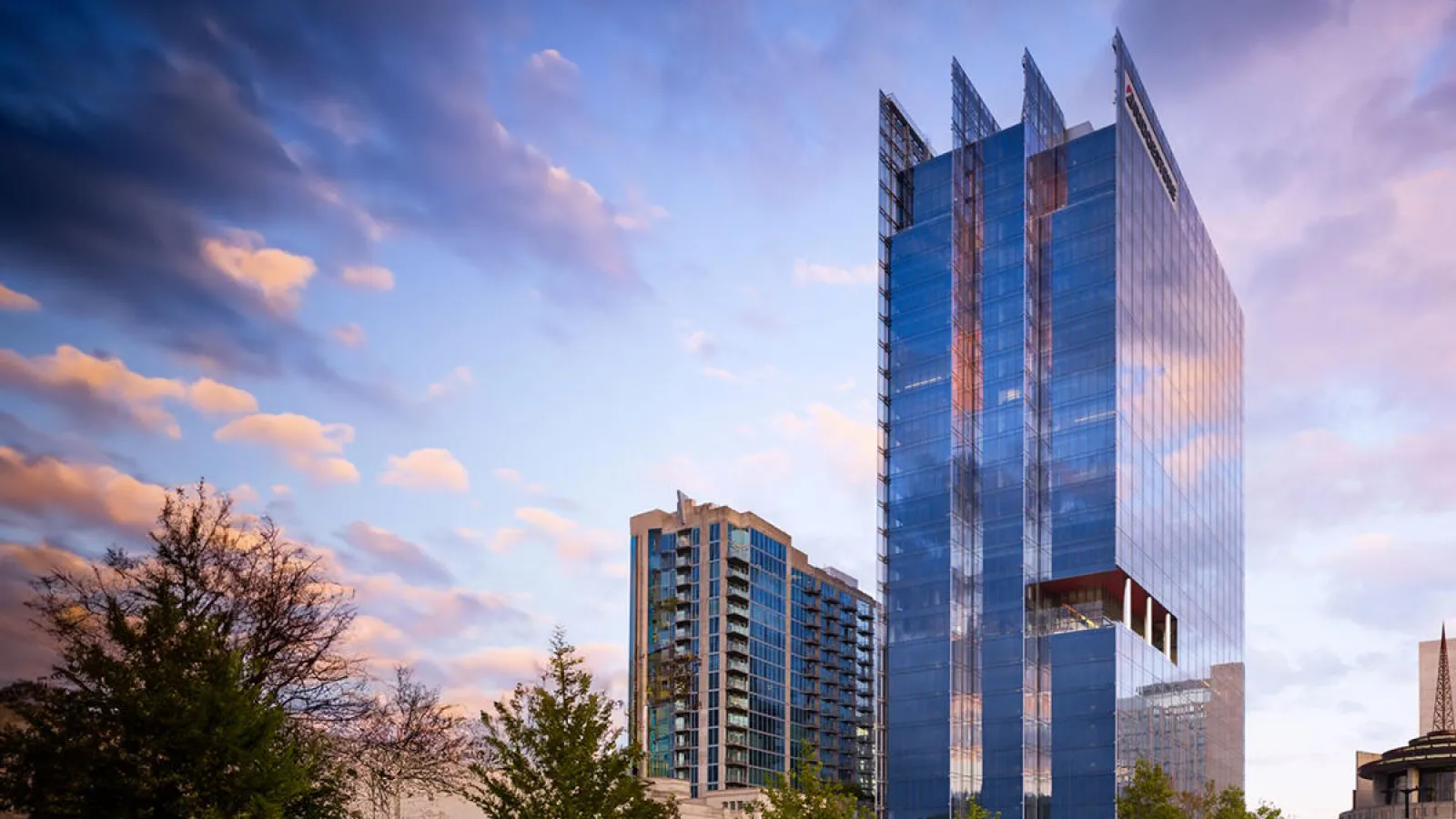 Modern glass skyscraper reflecting colorful sunset sky with surrounding trees and urban buildings.