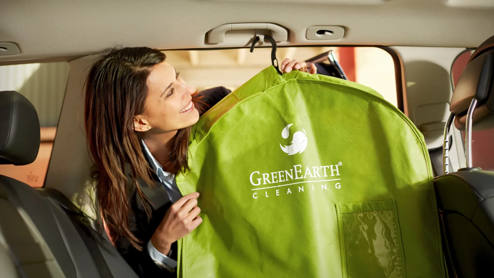 Woman in car holding a green garment bag labeled GreenEarth Cleaning, smiling and inspecting it.