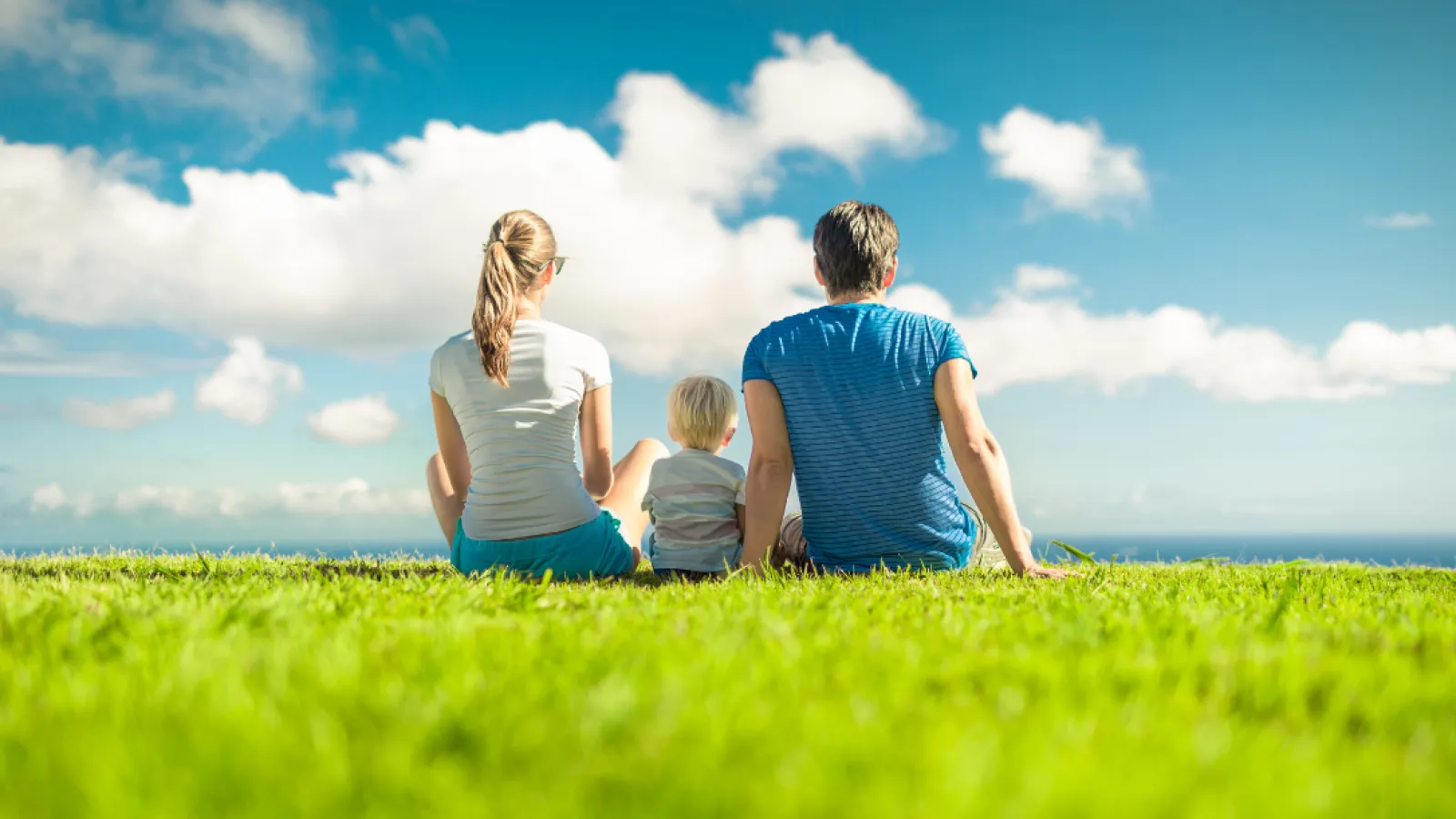 Family of three sitting on grass looking at blue sky with clouds on a sunny day.