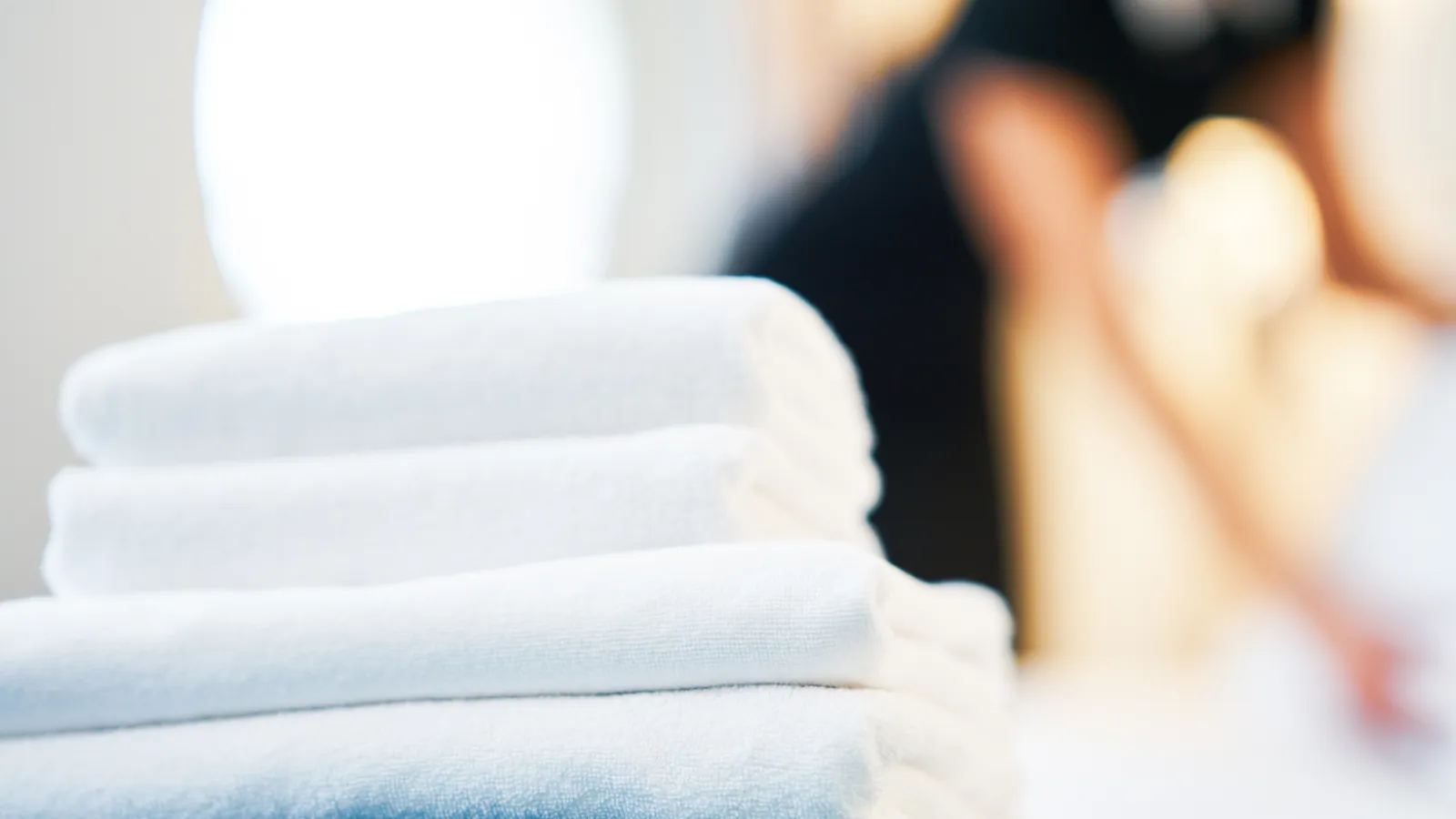 Stack of clean white towels in the foreground with a blurred person in the background setting a bed.