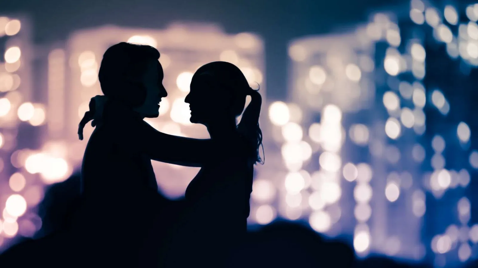 Silhouettes of a couple embracing against a blurred city lights background at night.