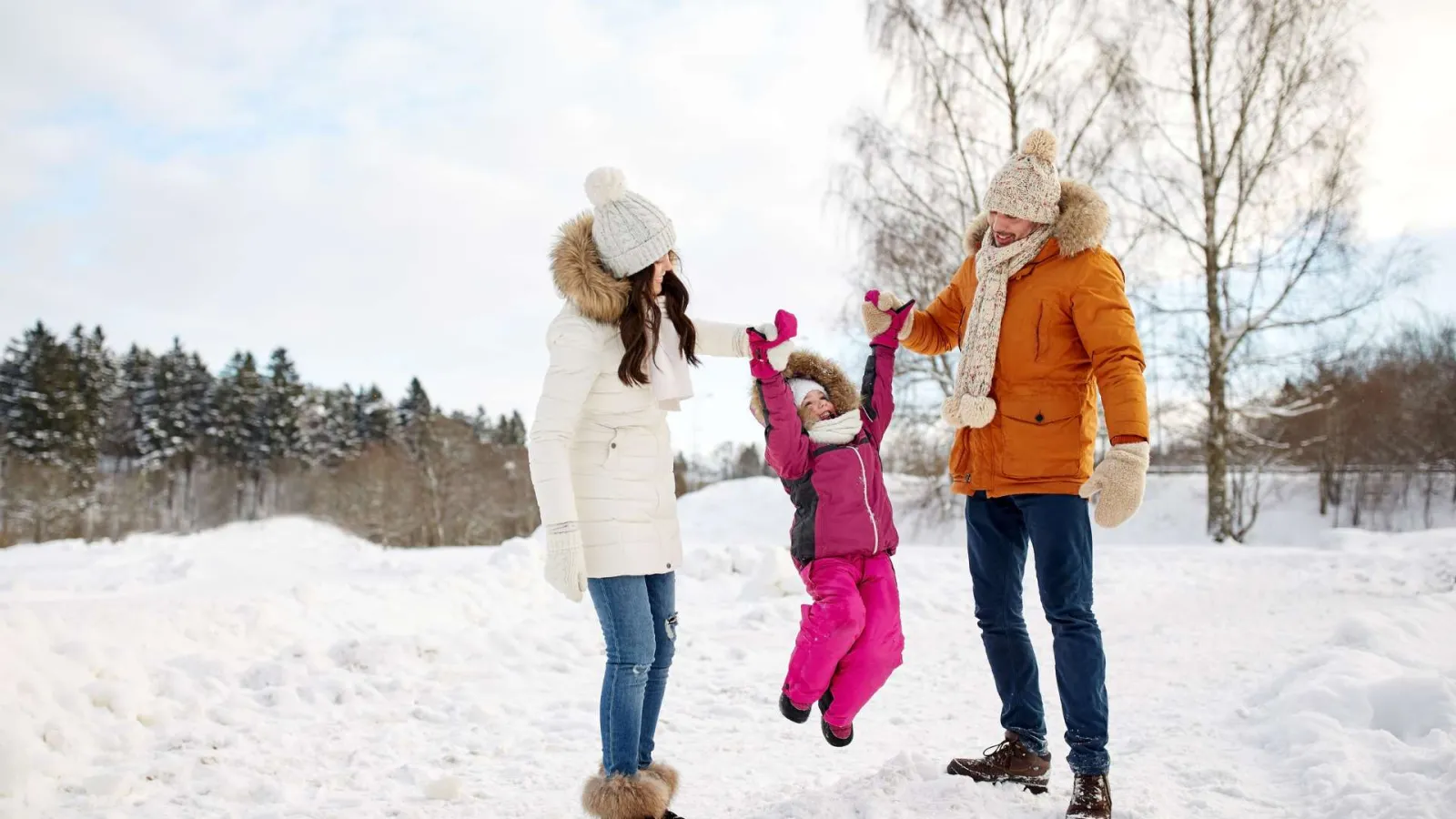 Parents in winter clothing lifting their child by hands outdoors in snowy landscape during daytime.