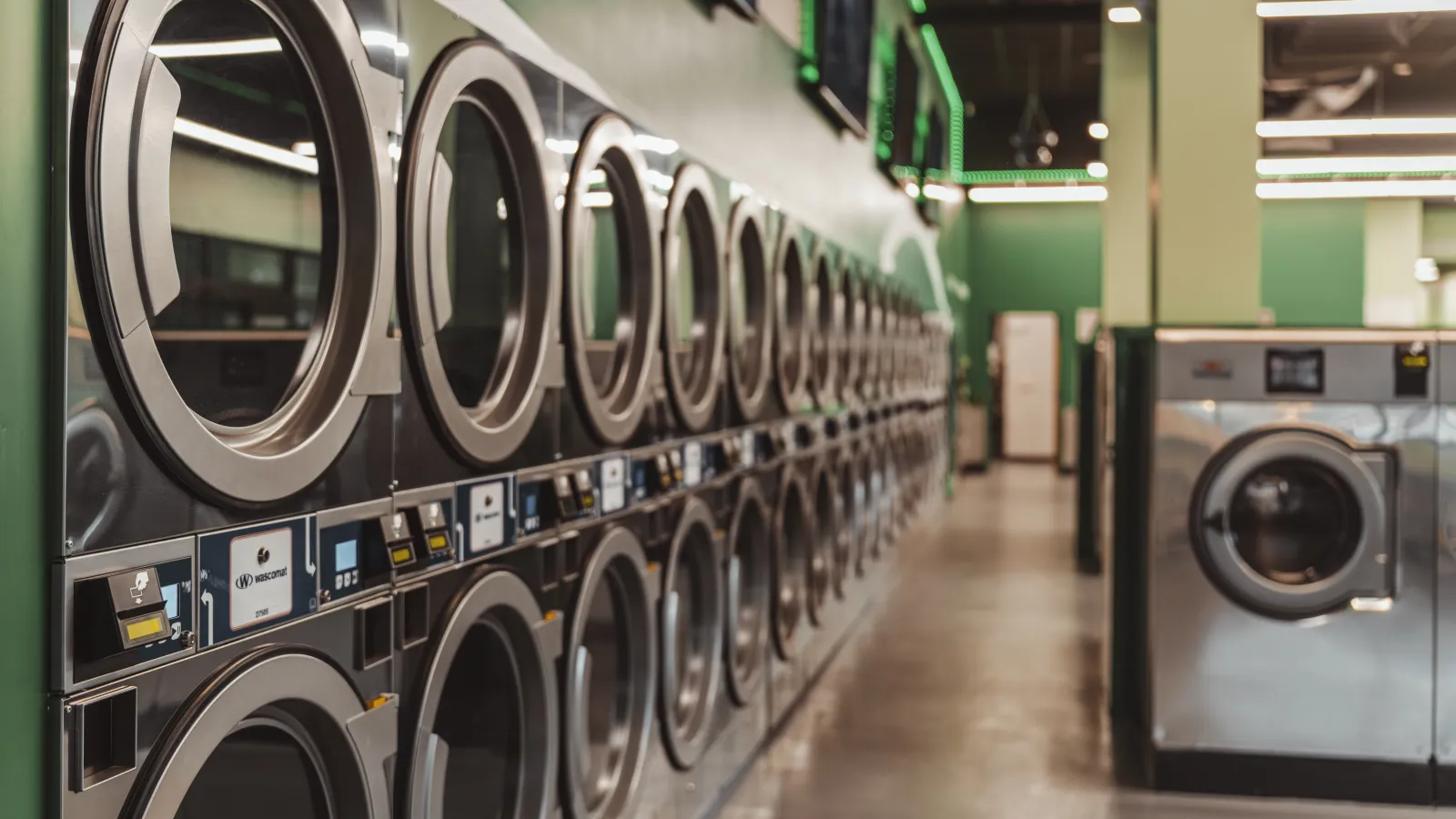 Row of modern stainless steel washing machines in a clean, well-lit laundromat with green accents.