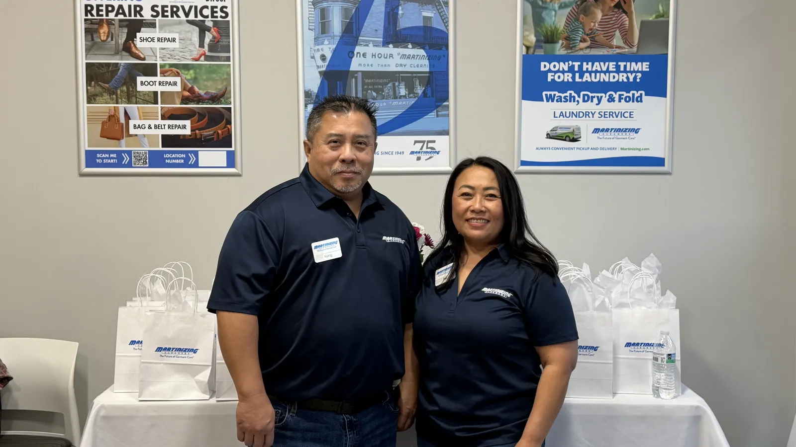 Two employees in navy shirts stand in front of a table with branded bags and repair and laundry service signs on a wall.