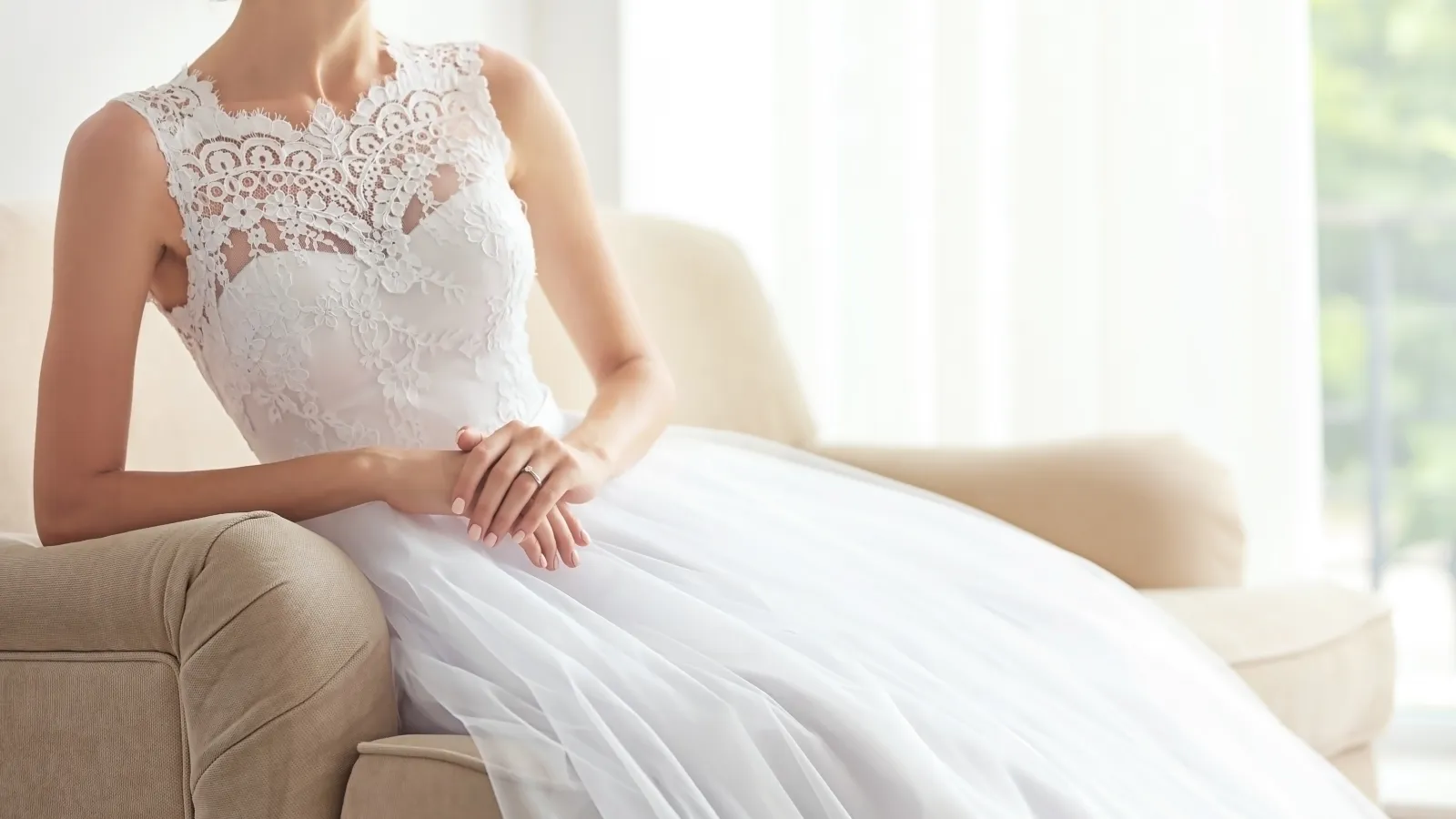 Bride in a white lace sleeveless wedding dress sitting on a beige armchair in natural light.