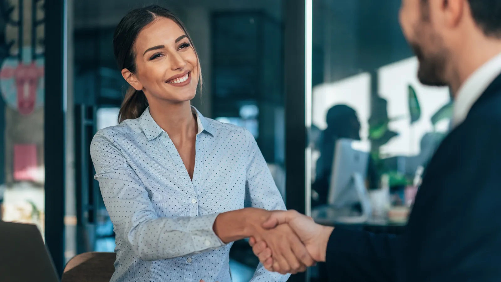 Businesswoman smiling and shaking hands with a colleague in a modern office setting.