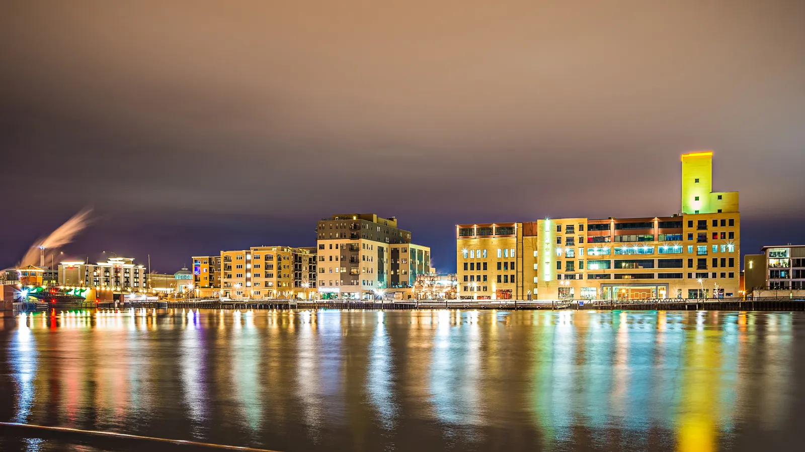 Nighttime cityscape with illuminated buildings reflecting on calm waterfront under cloudy sky.