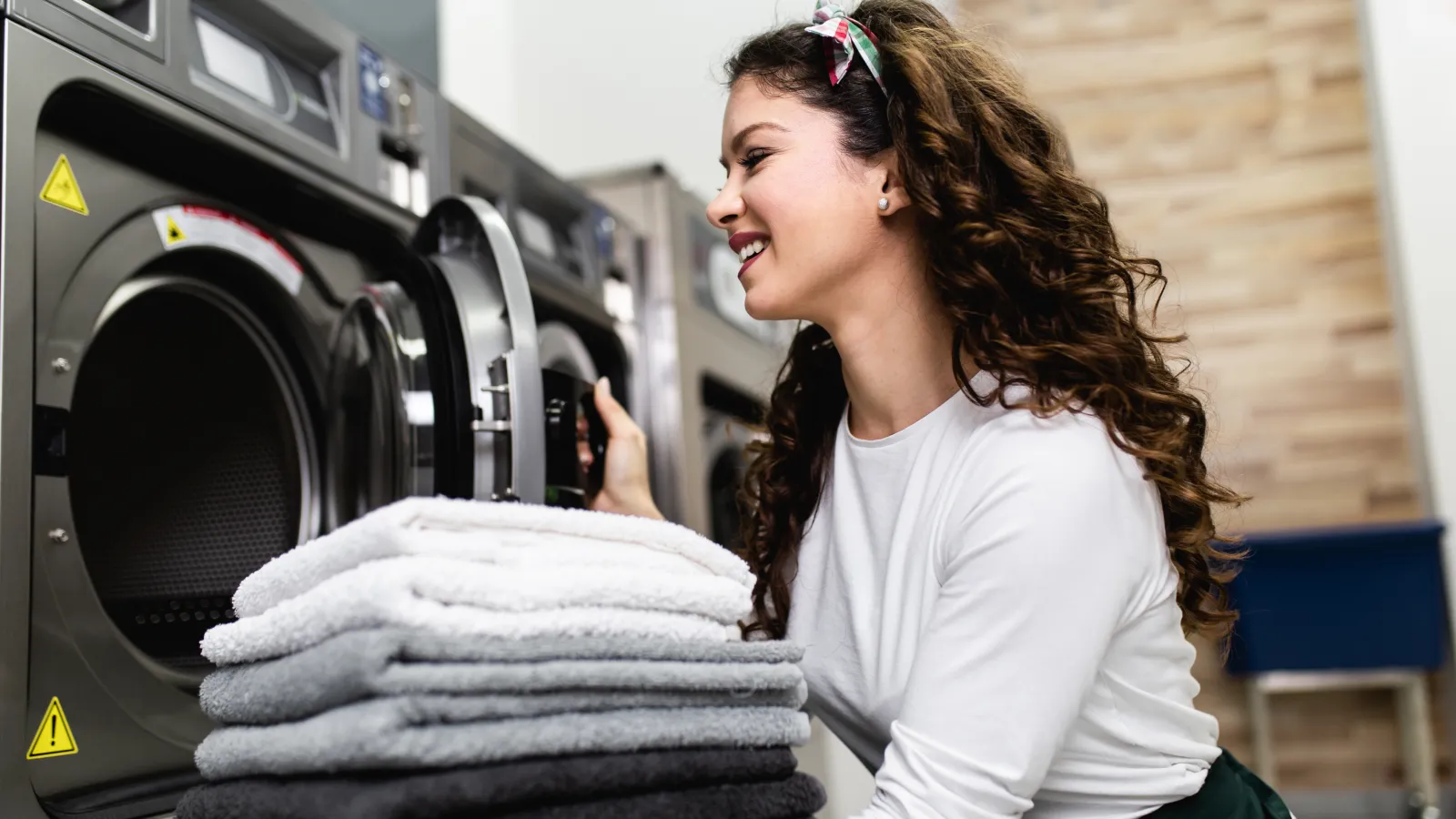 Smiling woman loading laundry into a washing machine with a stack of folded towels nearby in a modern laundry room.