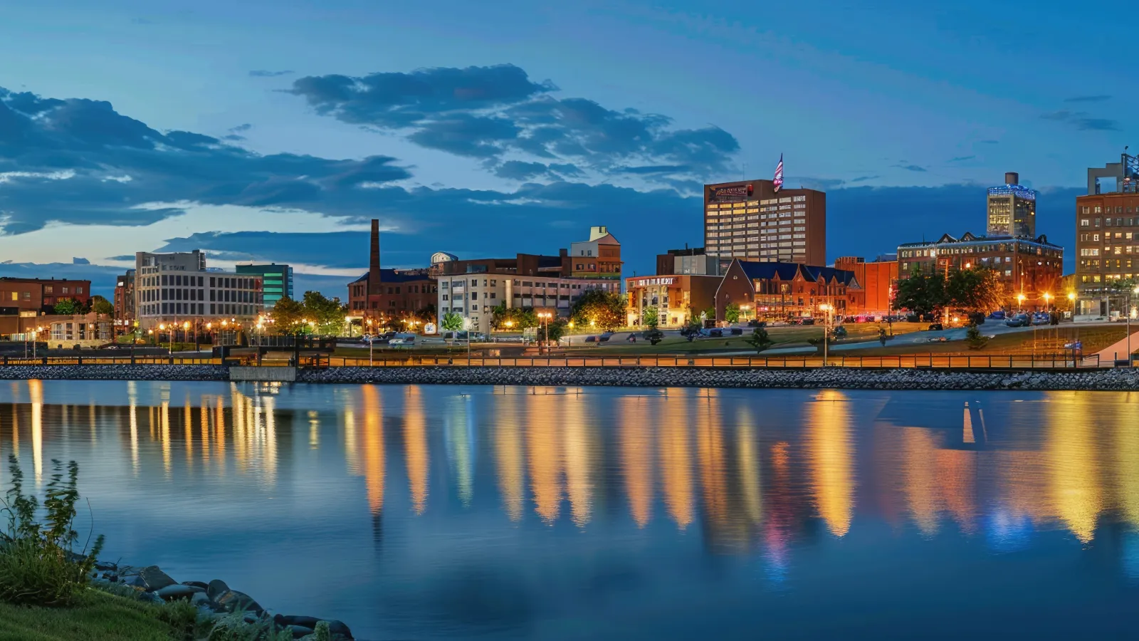 City skyline at dusk reflecting colorful lights on calm river water with clear blue evening sky.