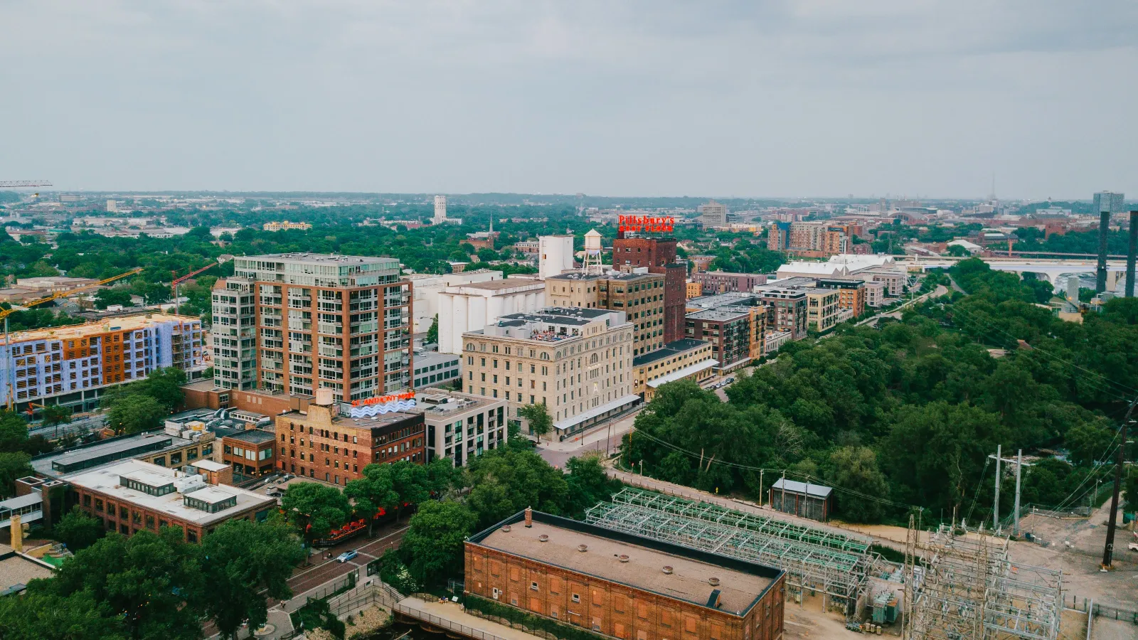 Aerial view of an urban cityscape with mixed-use buildings, dense trees, and a distant river under cloudy sky.