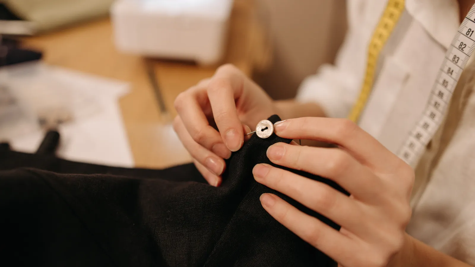Close-up of hands sewing a white button onto black fabric with a sewing needle and tape measure nearby