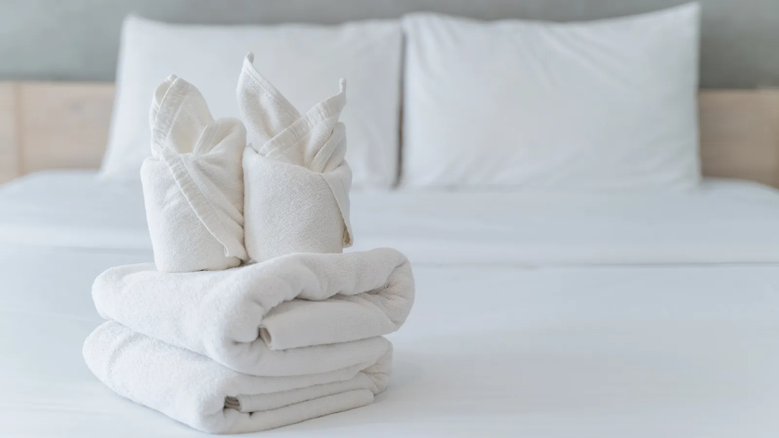 Stacked white towels with folded towel art on a neatly made white bed with pillows in background