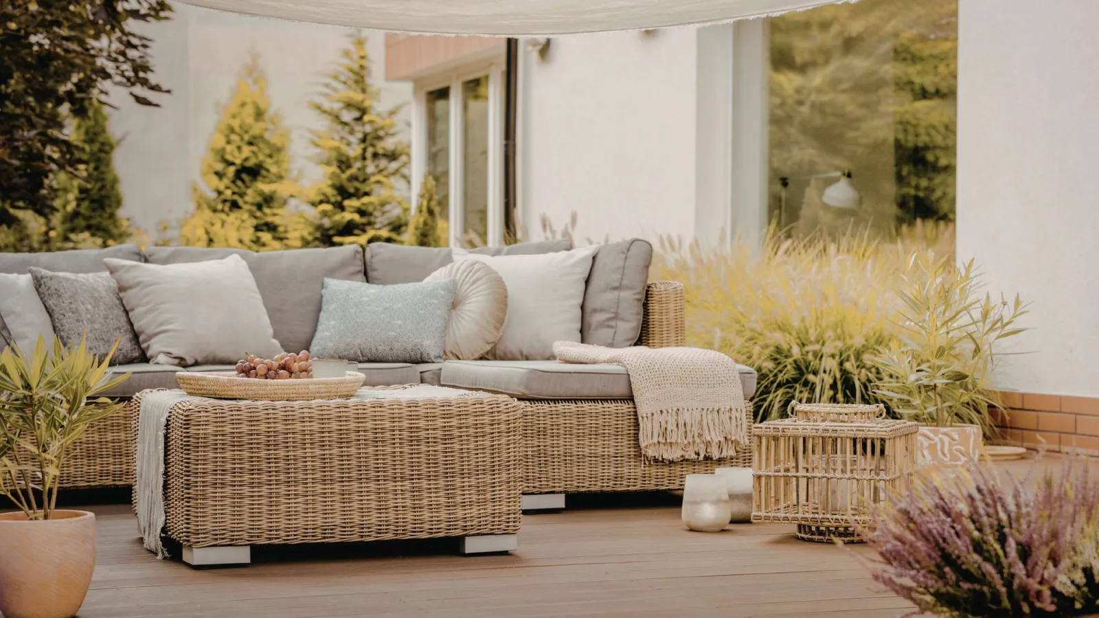 Cozy outdoor patio with wicker sofa, cushions, throw blanket, decorative plants, and wooden deck.