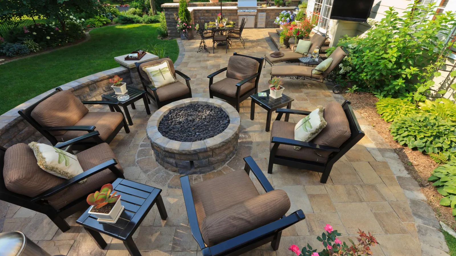 Outdoor patio with cushioned chairs arranged around a stone fire pit and lush garden landscaping.
