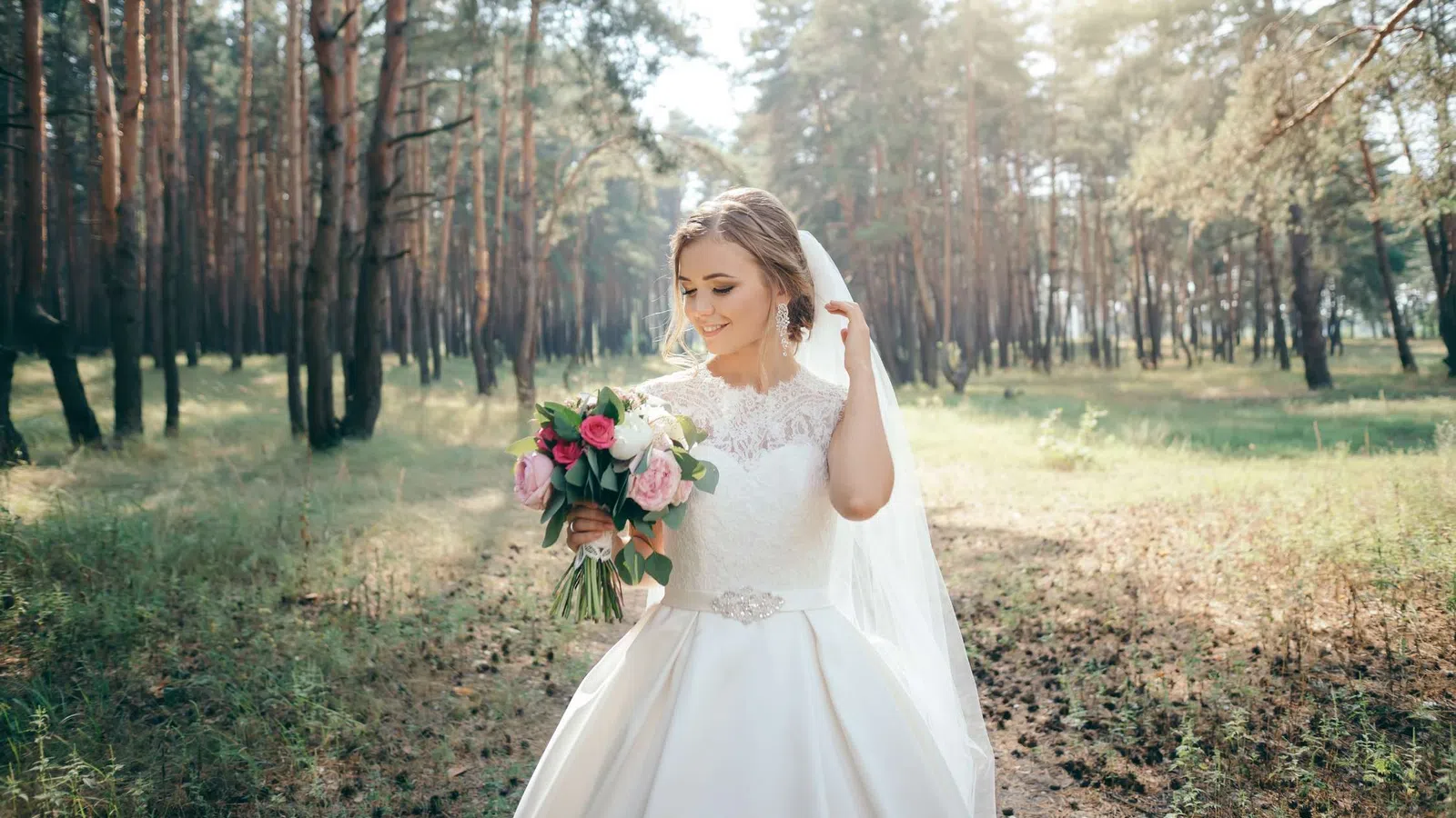 Smiling bride in white lace wedding dress holding pink and white bouquet in sunlit forest clearing