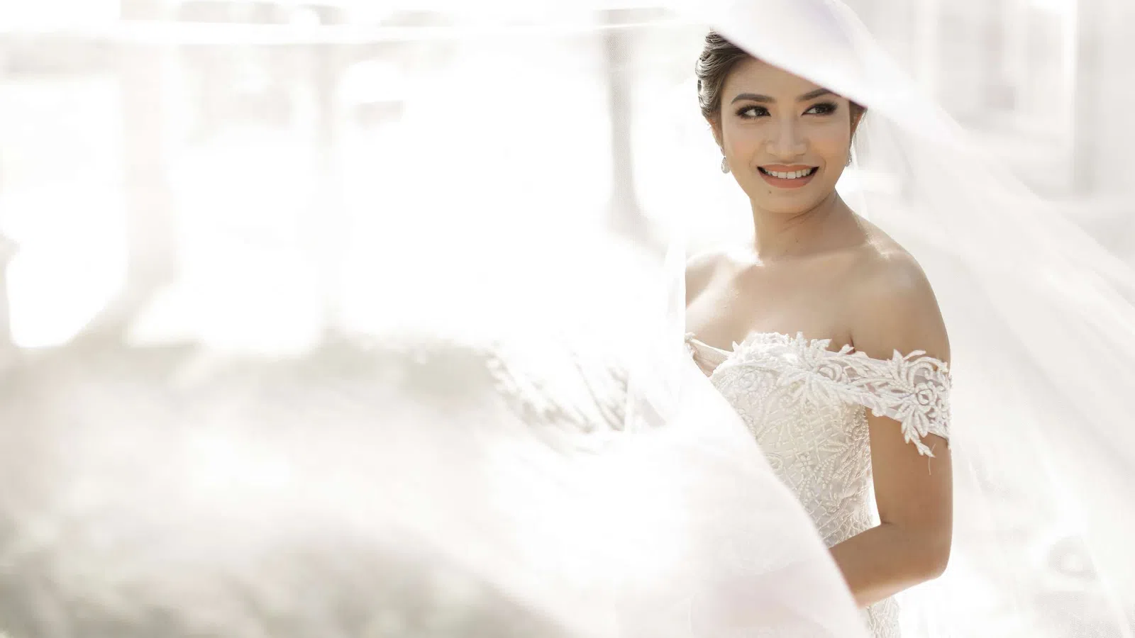 Smiling bride in a white lace off-shoulder wedding dress with flowing veil in soft natural light.