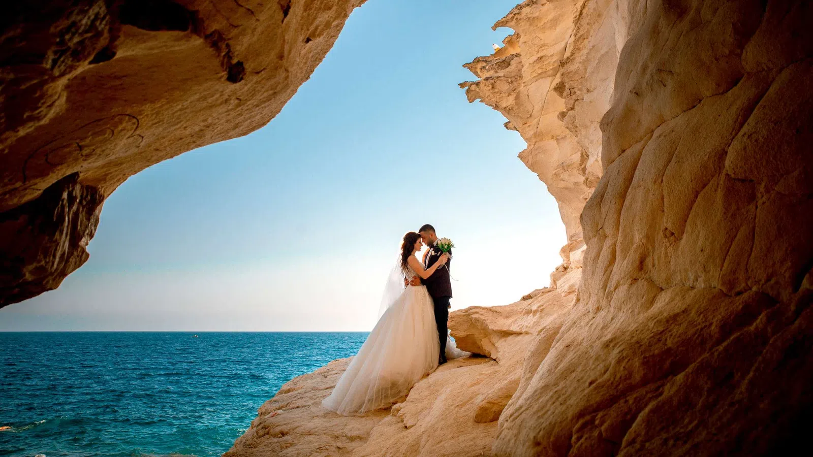 Bride and groom embracing on rocky cliff by the blue sea under clear sky during outdoor wedding photoshoot.