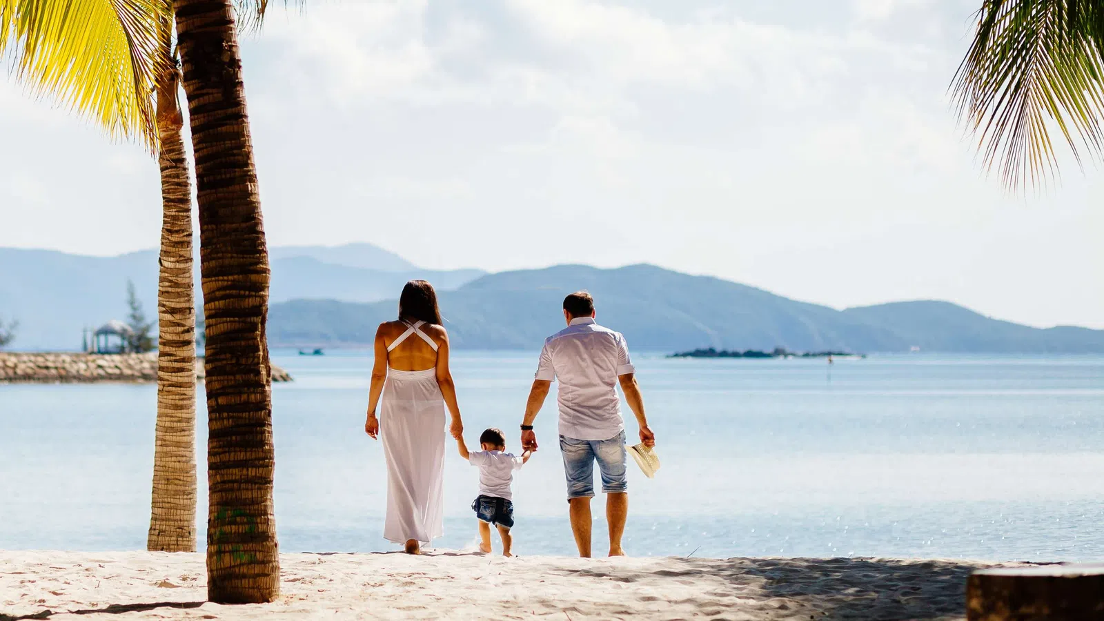 Family walking on sandy beach holding hands with ocean and mountains in the background under palm trees