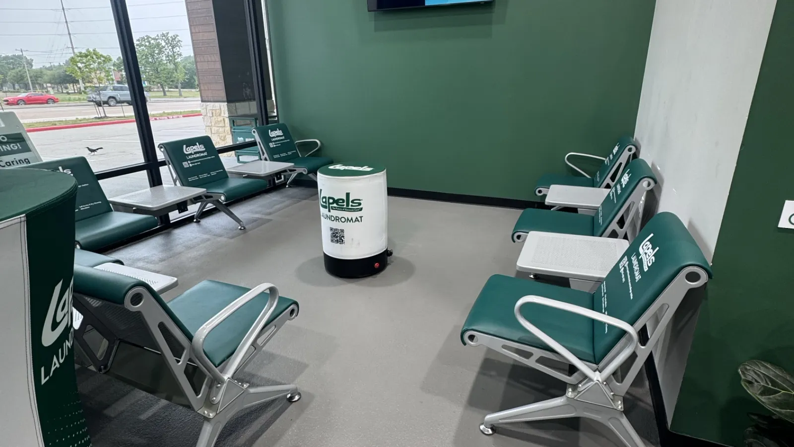 Waiting area inside a green-themed laundromat and dry cleaner with chairs, a TV, and a small round table.