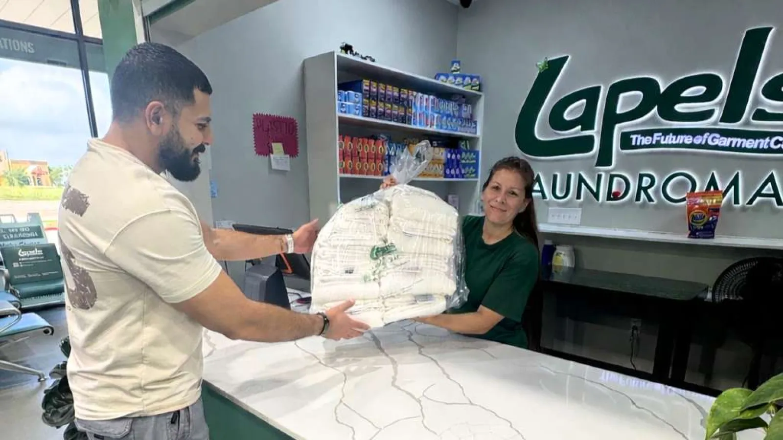 Man receives a large wrapped bundle of laundry from a staff member at Lapels Laundromat and Dry Cleaners counter.