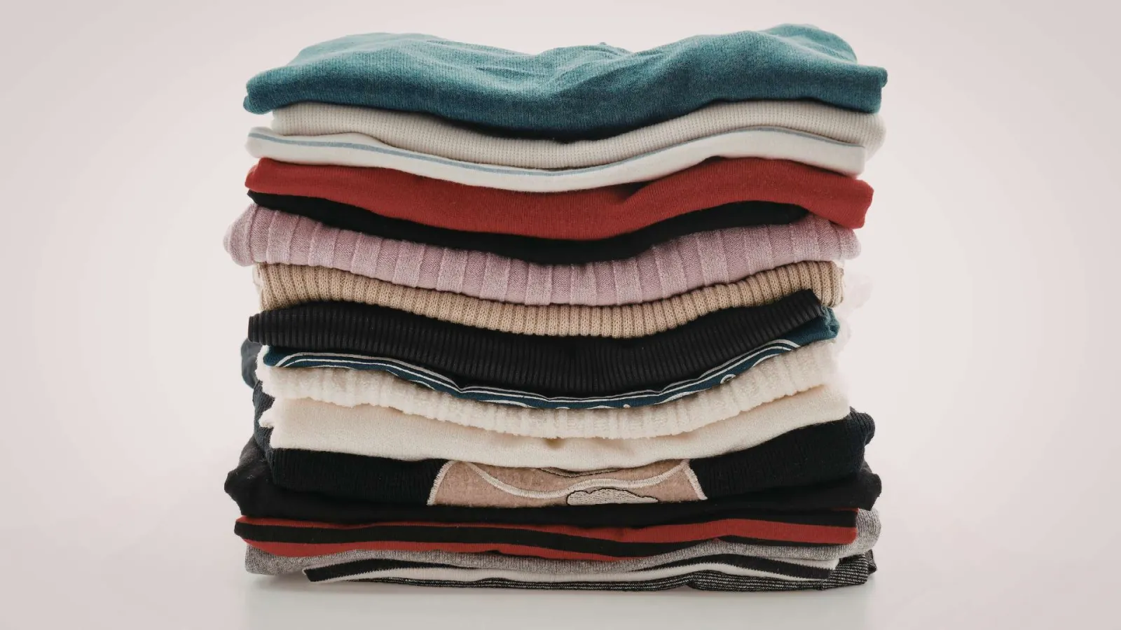 Stack of neatly folded colorful sweaters and knitwear on a plain background.