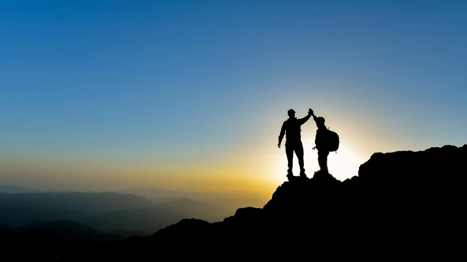 Two hikers high-fiving atop a mountain at sunset with a clear sky and distant mountain ranges.