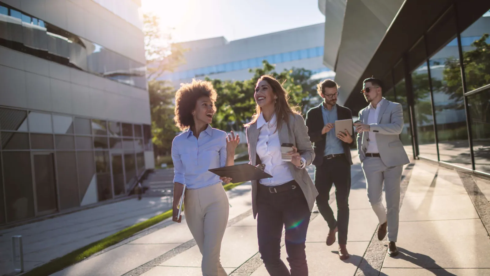 Four diverse business professionals walking and talking outside a modern office building on a sunny day.
