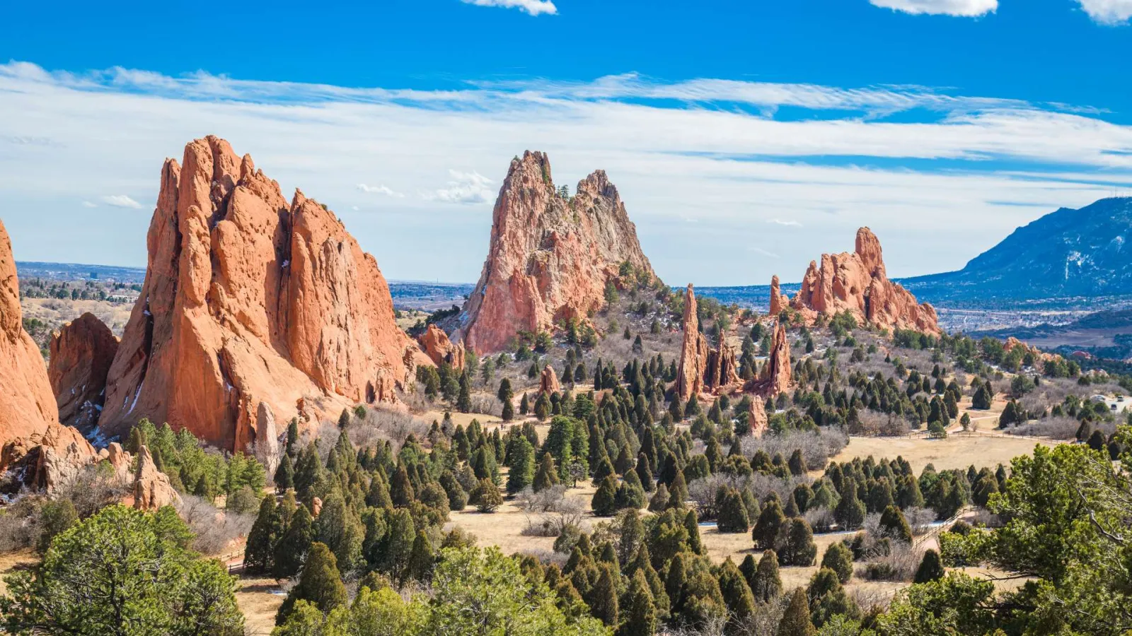 Red rock formations under blue sky with scattered clouds in a high desert landscape with green shrubs and trees.