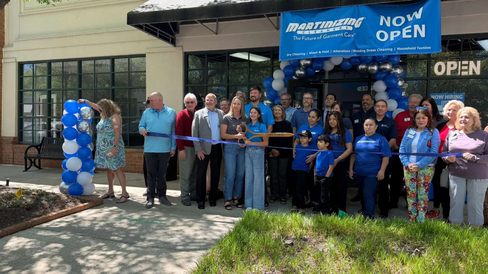 Group of people at Martinez Cleaners grand opening ceremony with blue balloons and ribbon cutting outdoors.