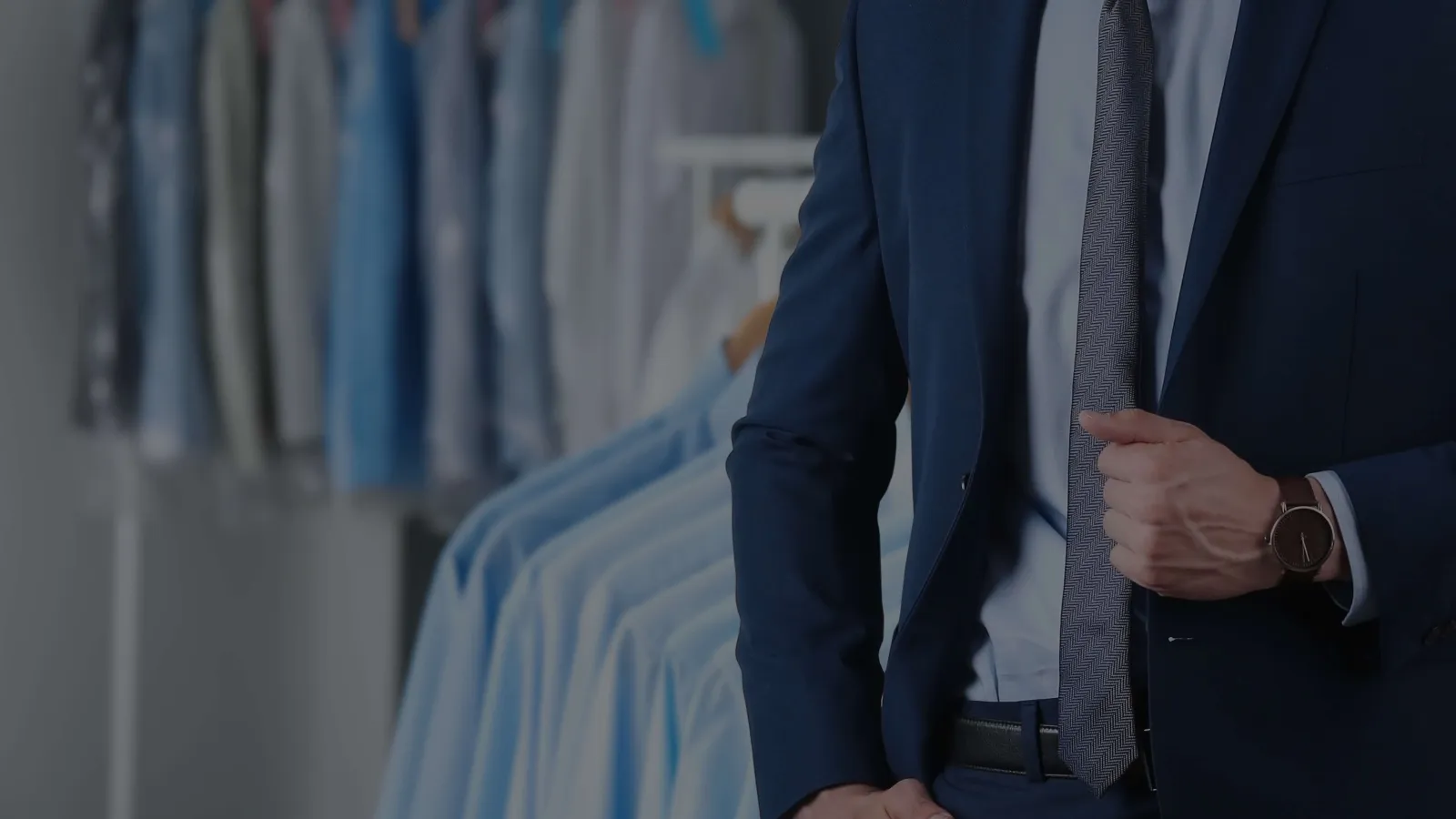 Man in navy suit adjusting tie in front of hanging light blue dress shirts in clothing store.