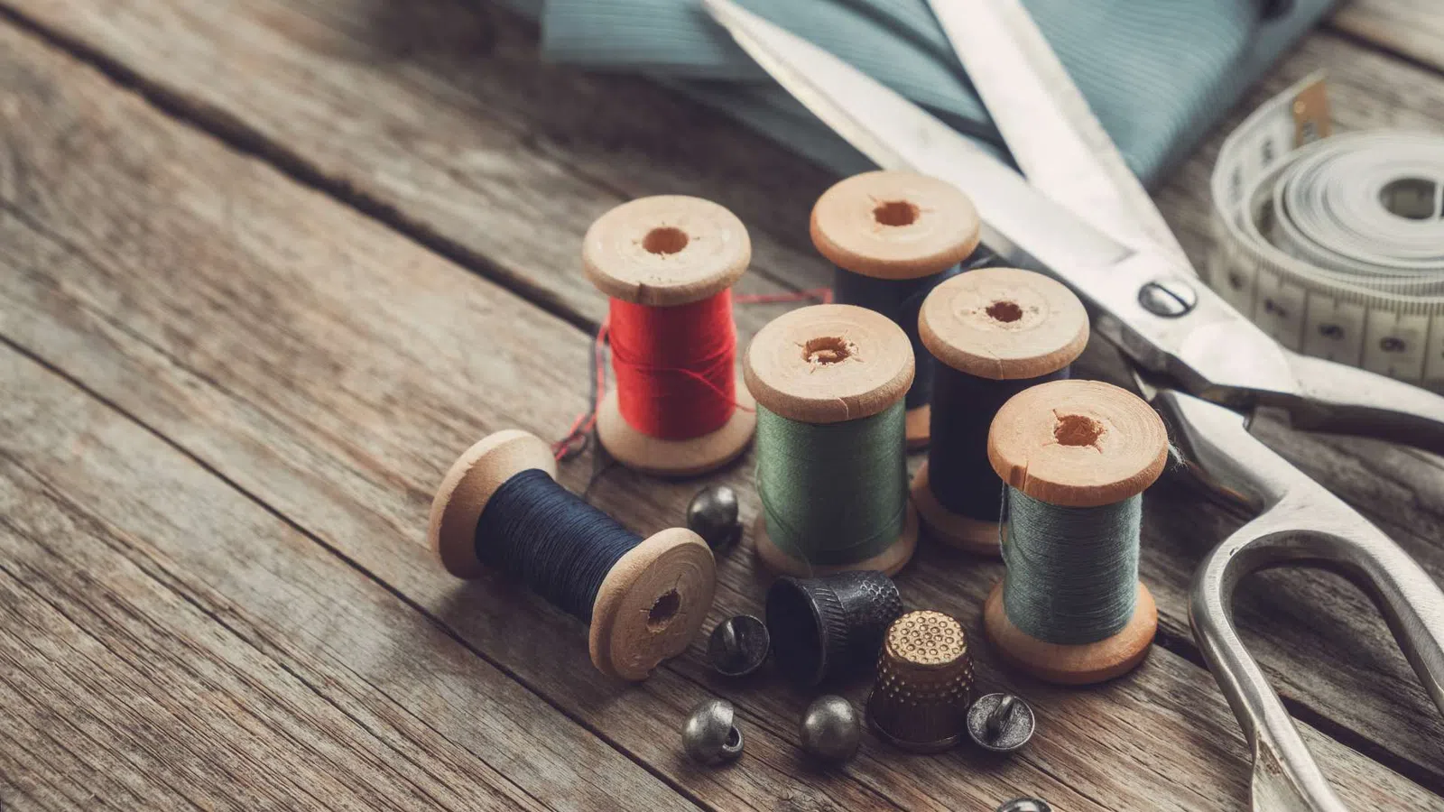 Vintage wooden spools of thread, sewing scissors, tape measure, thimbles, and sewing pins on rustic wood.