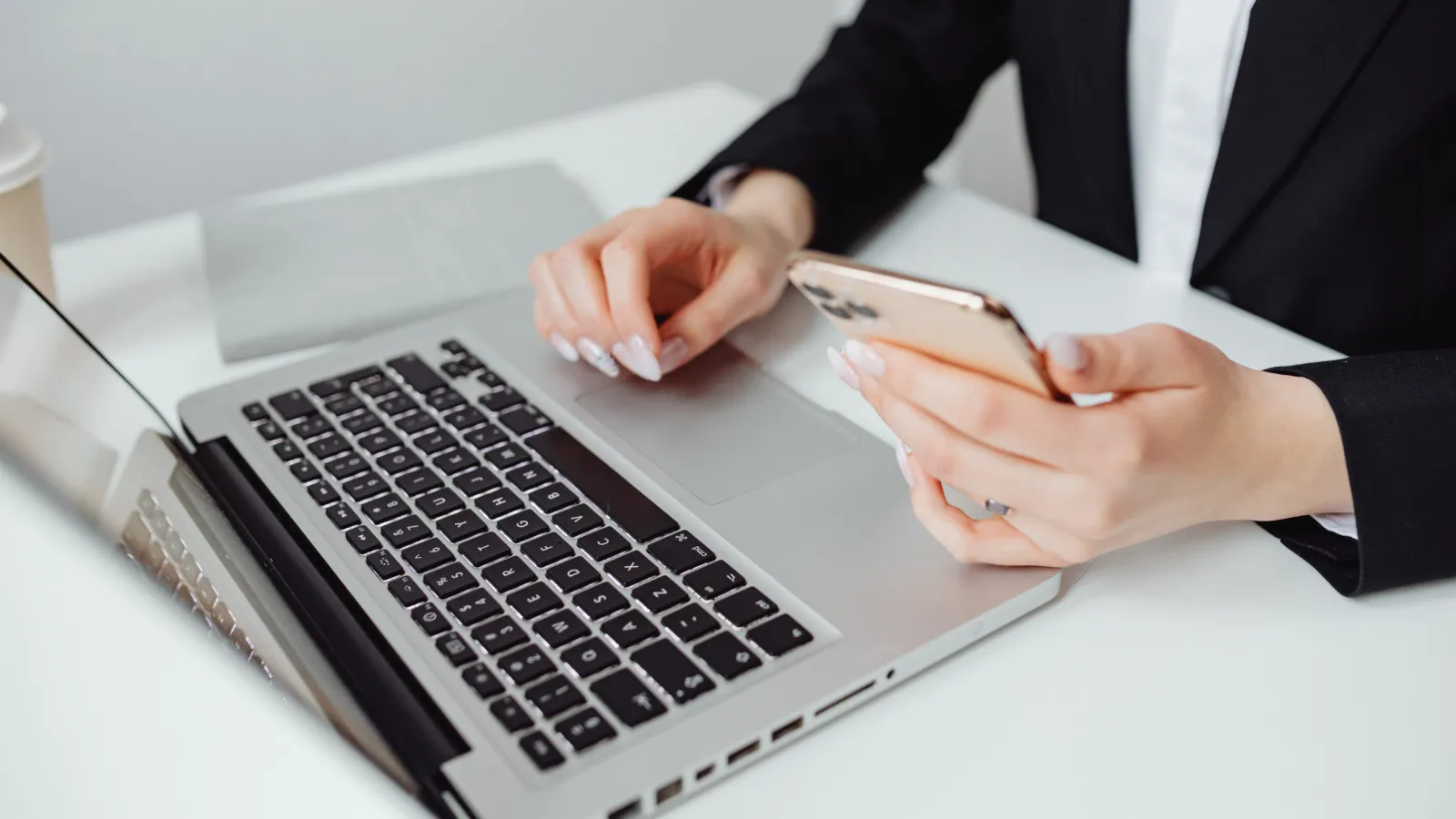 Person using smartphone and laptop on a white desk with a coffee cup nearby