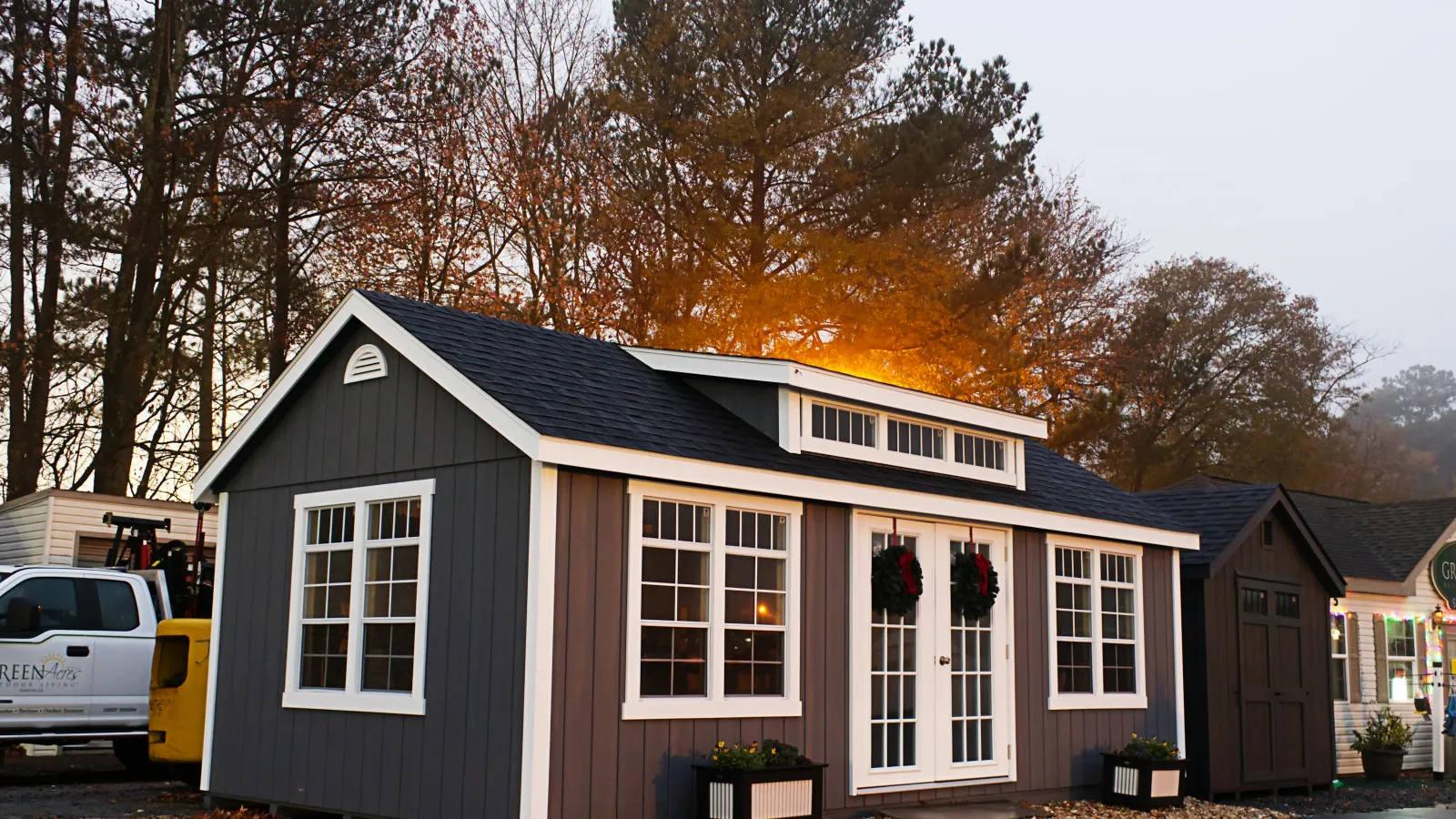 Modern gray garden shed with white trim and double doors decorated with wreaths, set against autumn trees at sunset.