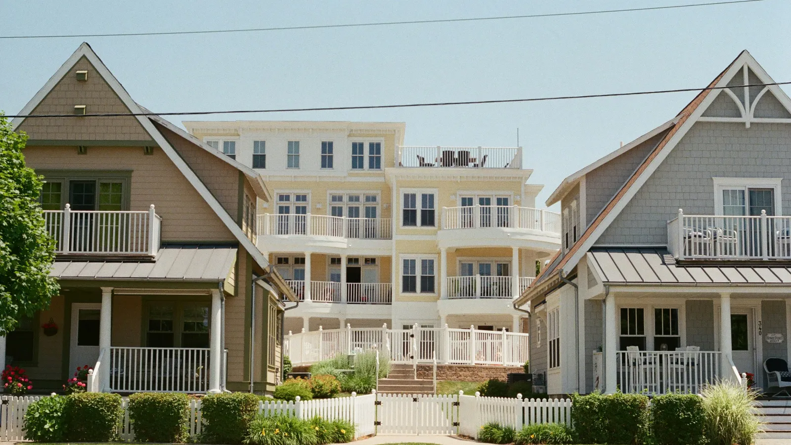 Two cozy suburban houses with porches and white fences flank a larger multi-story yellow building with balconies.