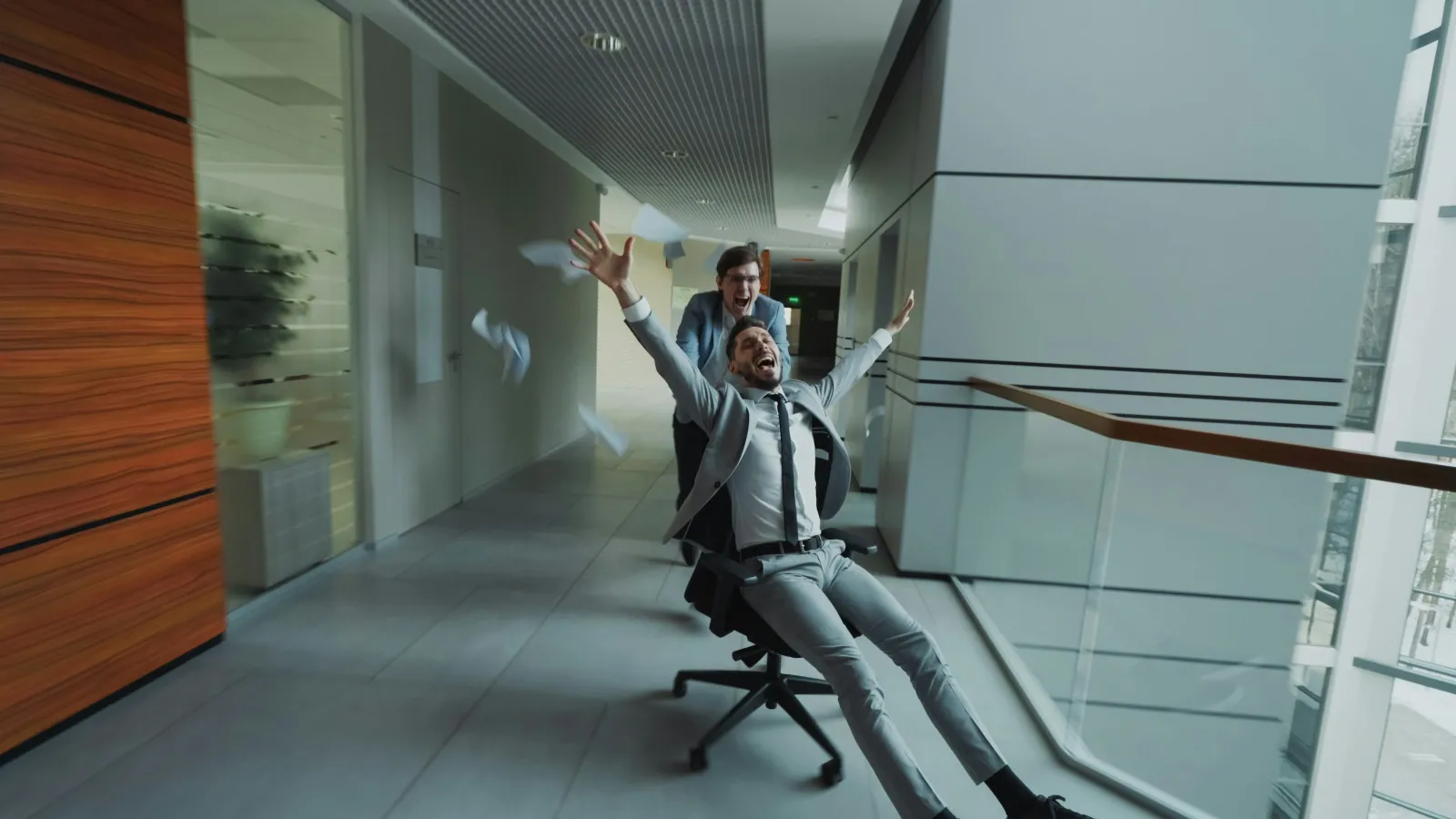 Two excited businessmen in suits riding an office chair down a modern office corridor, throwing papers in the air.