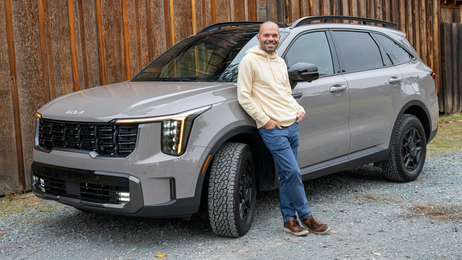 Man in beige hoodie and blue pants leaning against gray Kia SUV parked on gravel by wooden fence.